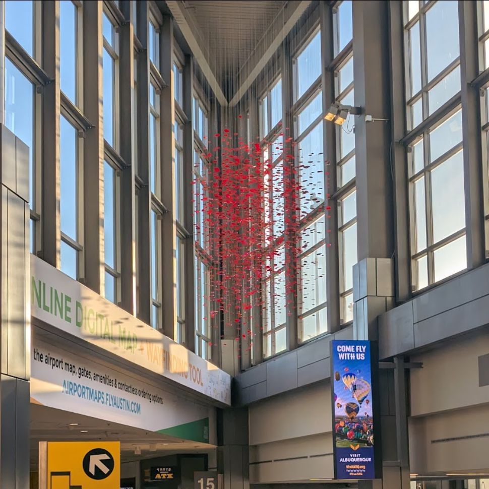 A large-scale red hanging art installation, made up of hundreds of suspended elements, is displayed in the high bay triangle area near Gate 15 inside Austin-Bergstrom International Airport. The installation is positioned against tall glass windows with natural light streaming in, while travelers move through the busy concourse below.